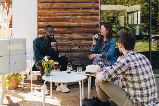 Amis réunis autour d'une table pour un apéritif sur terrasse en bois avec baie vitrée à côté d'un barbecue marcel gris soie